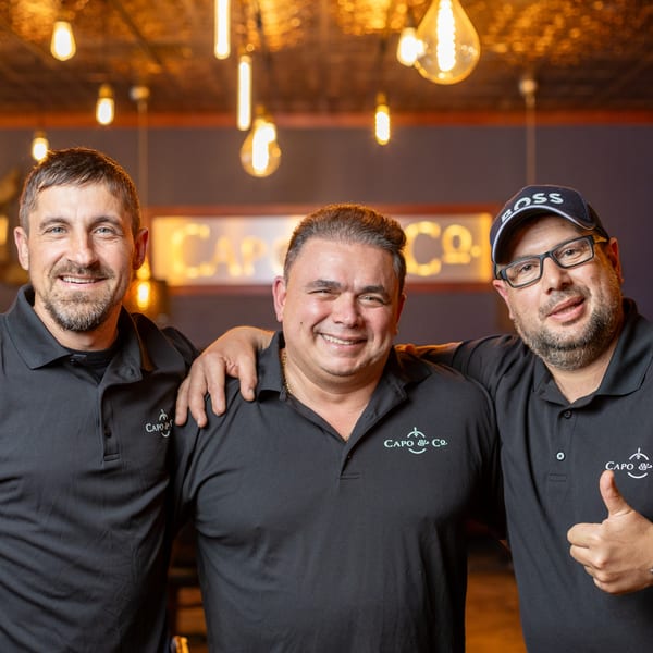 Three smiling men in black Capo & Co. shirts, one giving a thumbs up, in a warmly lit restaurant.