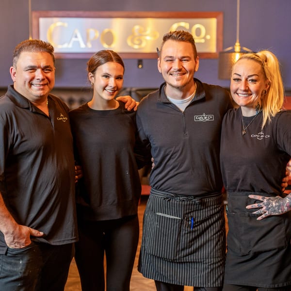 Four smiling Capo & Co. staff members, two men and two women, standing together in front of a sign.
