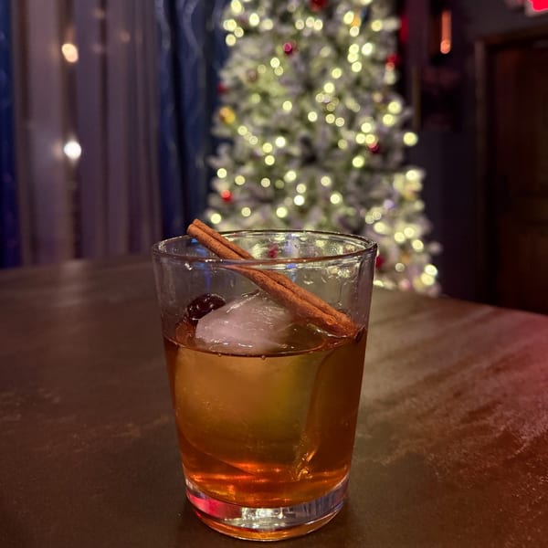 A Capo & Co. cocktail with cinnamon sticks and a cherry, in front of a blurred Christmas tree.