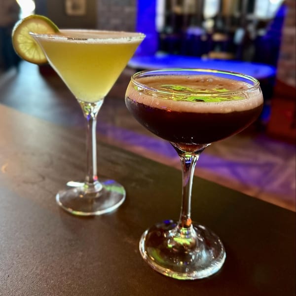 Two cocktails, one yellow with a lime, one dark with foam, on a bar at Capo & Co.