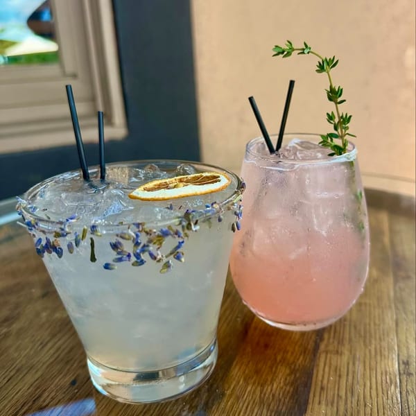 Two cocktails on a wooden table, one garnished with lavender and a dried orange, the other with thyme.