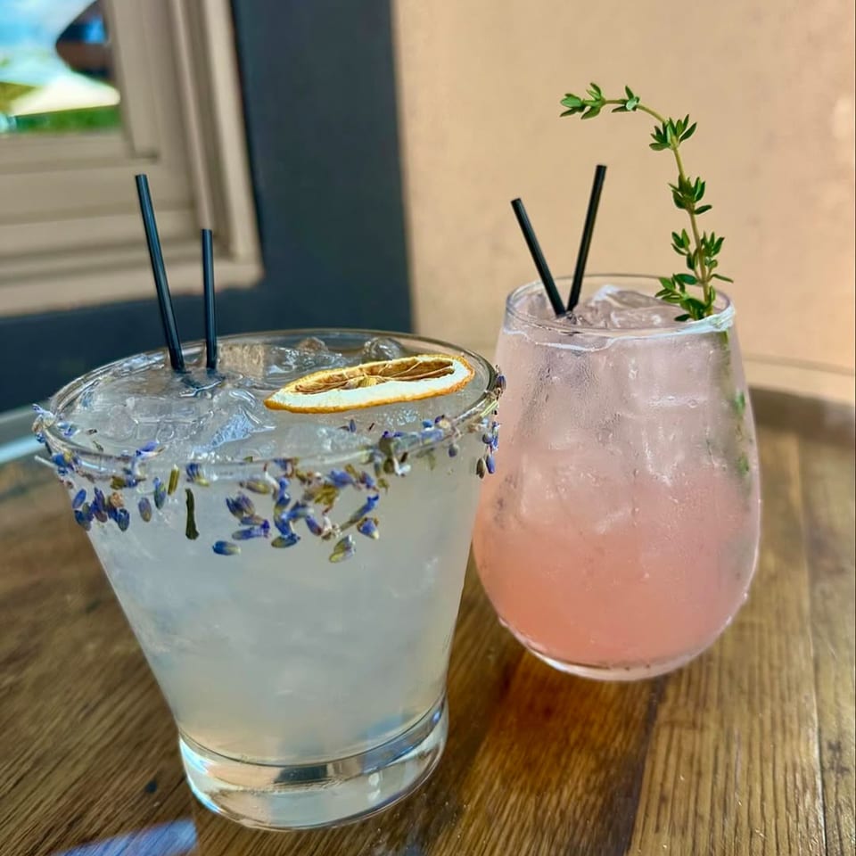 Two cocktails on a wooden table, one garnished with lavender and a dried orange, the other with thyme.