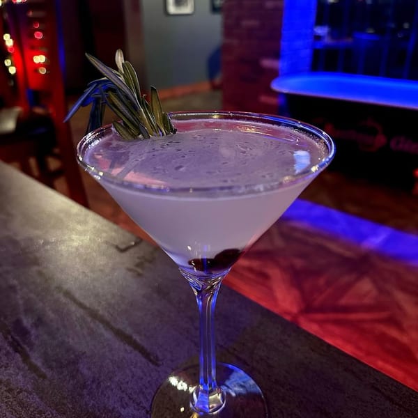 A martini glass with a light purple drink, garnished with rosemary and dark berries.