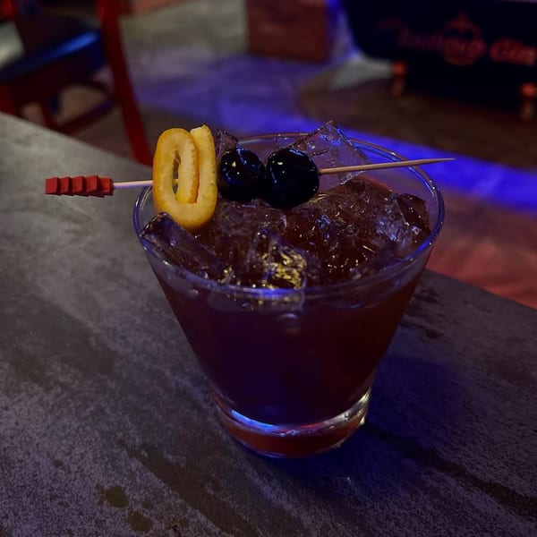 A dark red cocktail with ice, garnished with two cherries and an orange peel, on a dark surface.