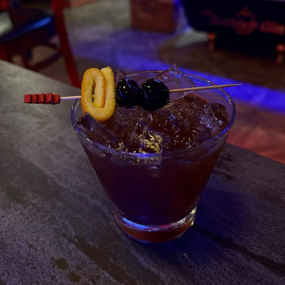 A dark red cocktail with ice, garnished with two cherries and an orange peel, on a dark surface.