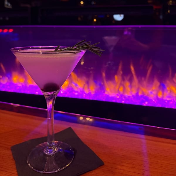 A martini with rosemary and a cherry sits on a bar with a purple and orange fireplace behind it.