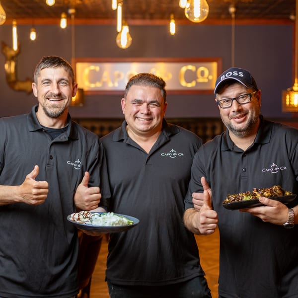 Three smiling men in black Capo & Co. shirts, holding food and giving thumbs up.