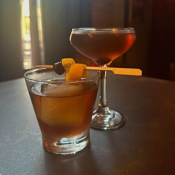 Two cocktails, one garnished with orange peel and cherry, on a dark table at Capo & Co.