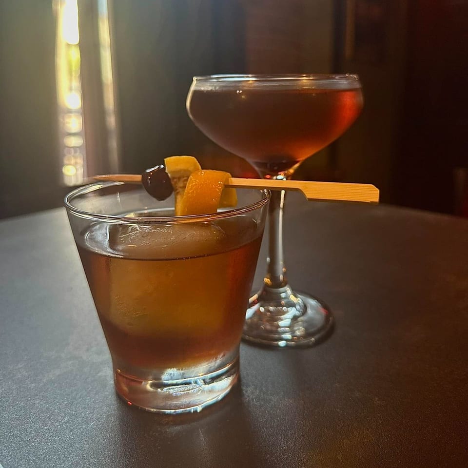 Two cocktails, one garnished with orange peel and cherry, on a dark table at Capo & Co.