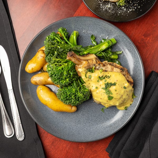 Grilled pork chop with mustard sauce, roasted potatoes, and broccolini on a gray plate.